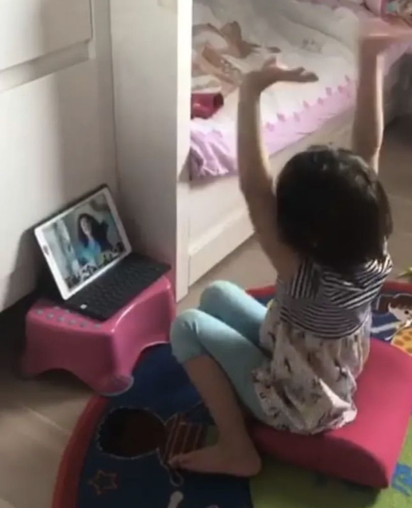 Real Diluu class: little girl singing the hello song with her arms up in front of her teacher on the tablet